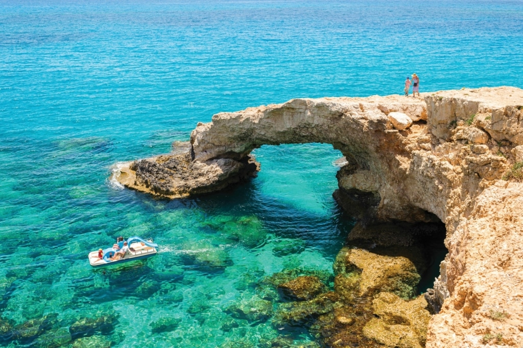 A rock formation on the ocean, with an arch. A small boat is in the sea and two people are on the rock.