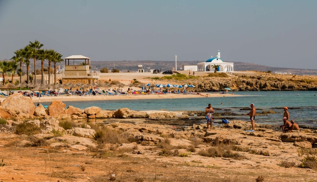 Land with some rocks by the sea. In the background there is a building with a blue roof. 