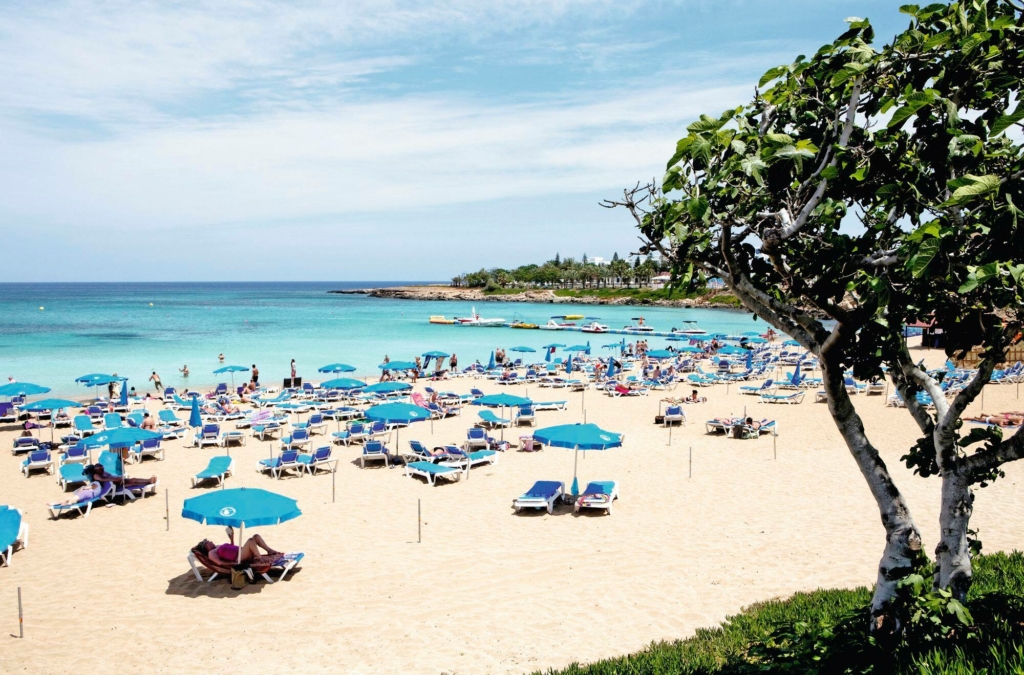 A beach scattered with sunbeds and umbrellas. A tree is in the foreground and the sea is in the background.