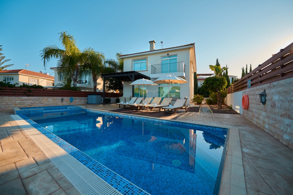 The back garden of a villa, with a swimming pool, palm tree and deckchairs.