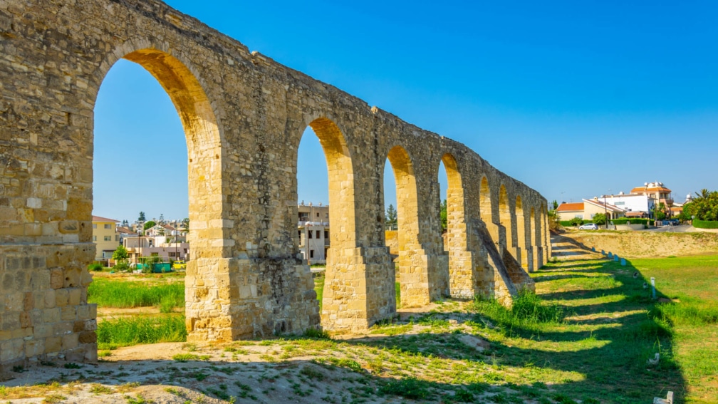 An old wall with arches, surrounding it is a grassy field and buildings in the background.