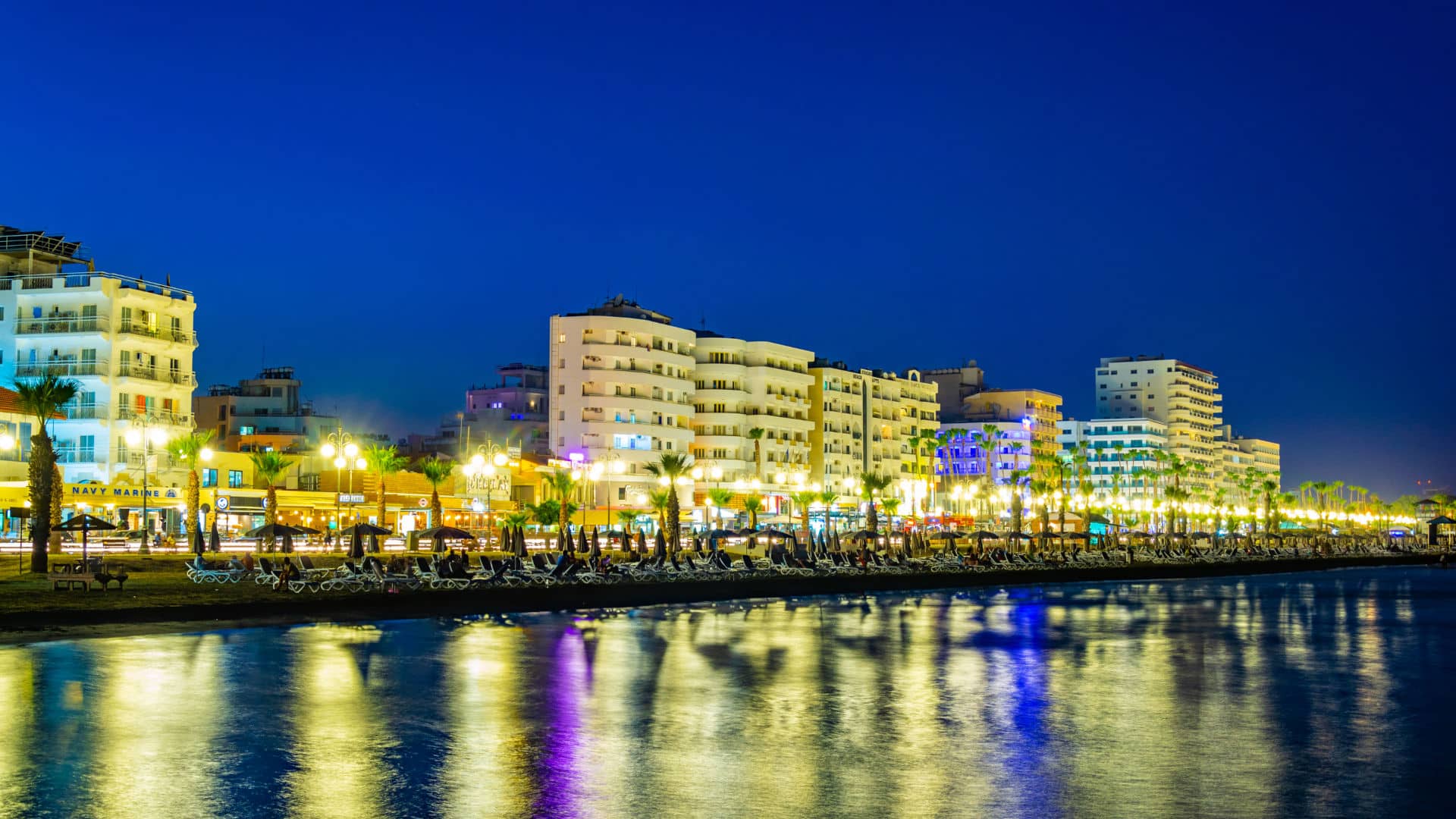 A photo of Larnaca at night time, with buildings and street lights in front a beach with deckchairs.