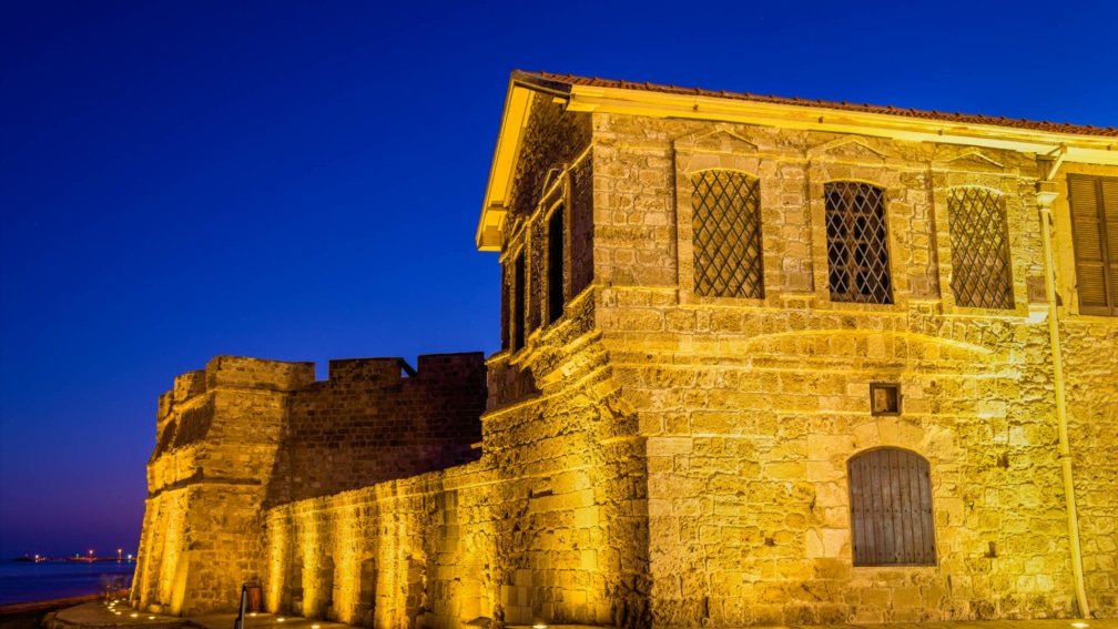 An old building at night, lit up by streelights. The sea is in the background.