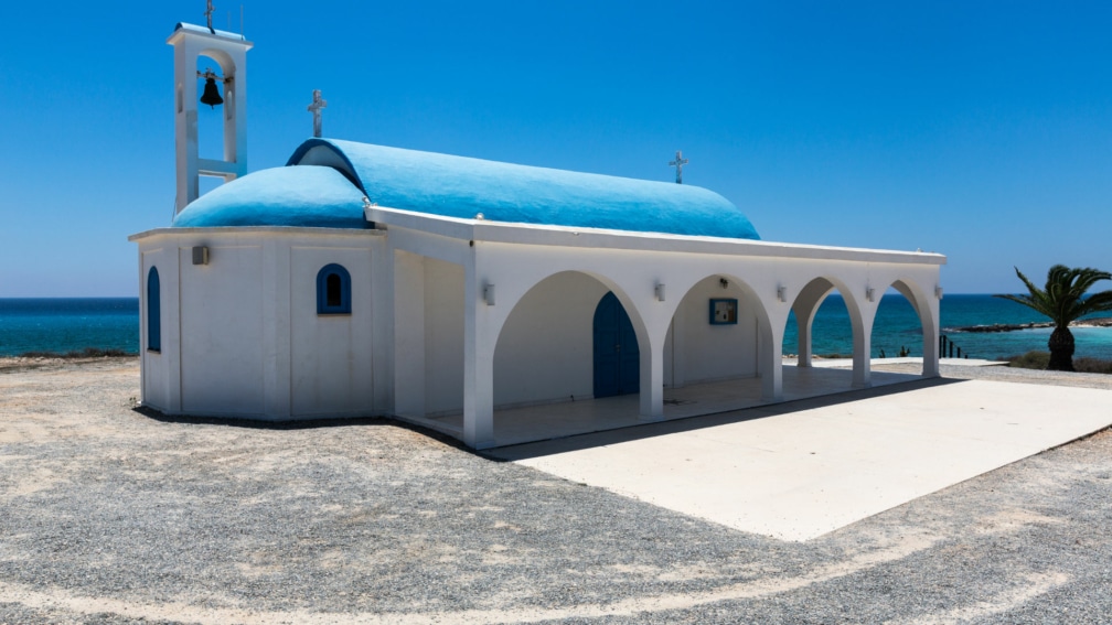 A church with a blue roof, and a bell on the side. In the background there is a plam tree and the sea.