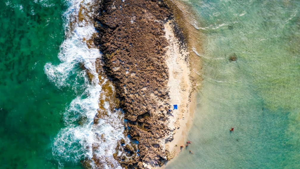 a birds eye view photo of the sea, with a large rock in the middle and a small beach.