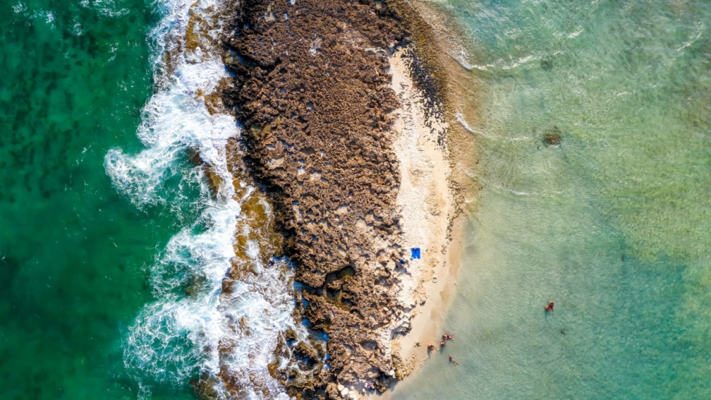 a birds eye view photo of the sea, with a large rock in the middle and a small beach.