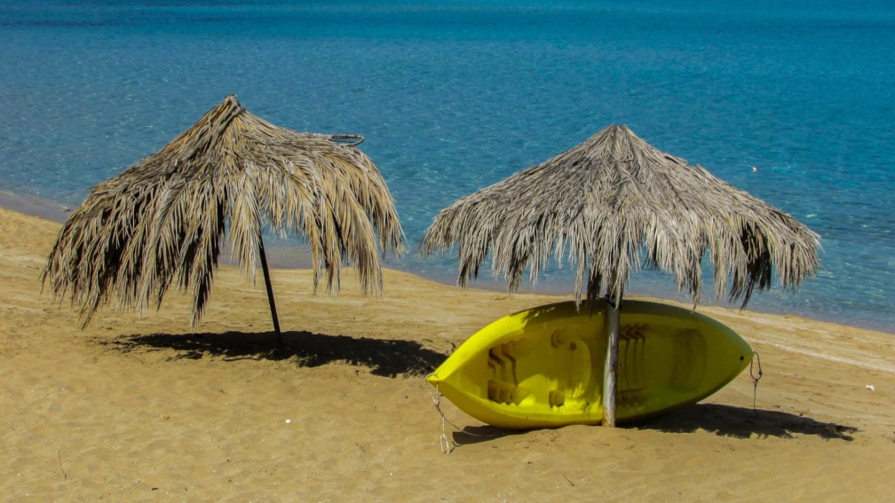 A sandy beach with two umbrellas made from leaves. A boat is underneath one of them in the shade.