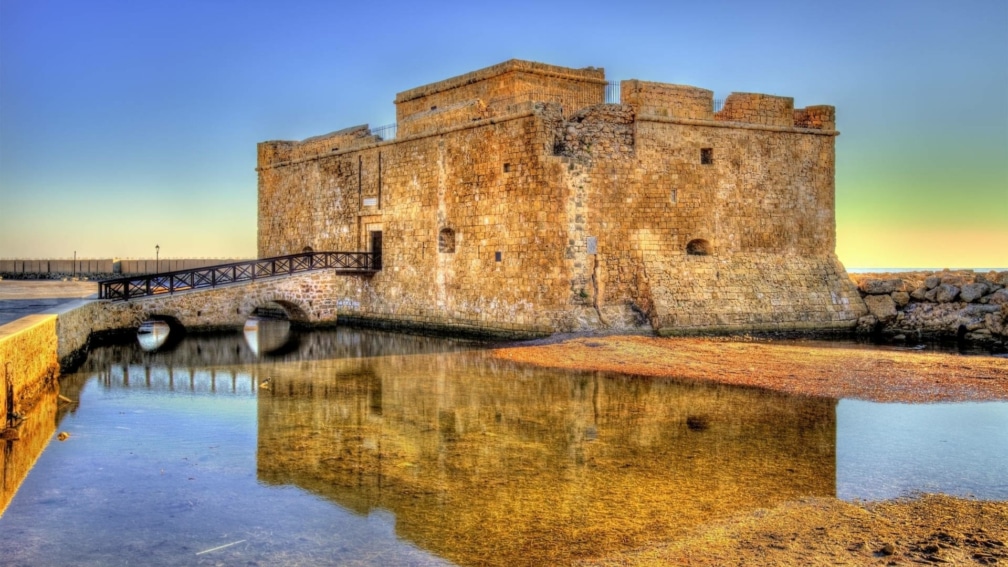 A small bridge leading to an old building on the water. The sea is in the background.