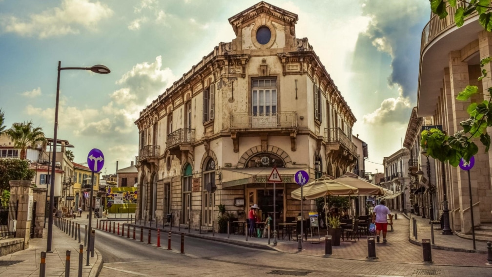 A street with an old building in the centre. Chairs and umbrellas are outside the building.