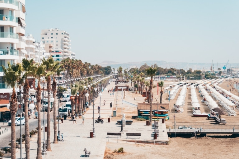 A city centre, with a walkway, palm trees, a beach and buildings.