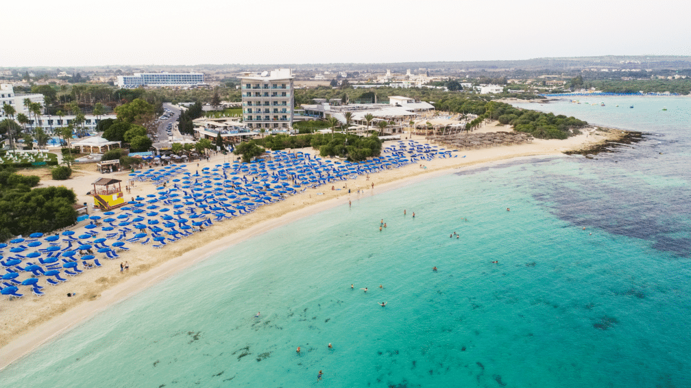 A high angle photo of the ocean, the beach with deckchairs on, and buildings in the background.