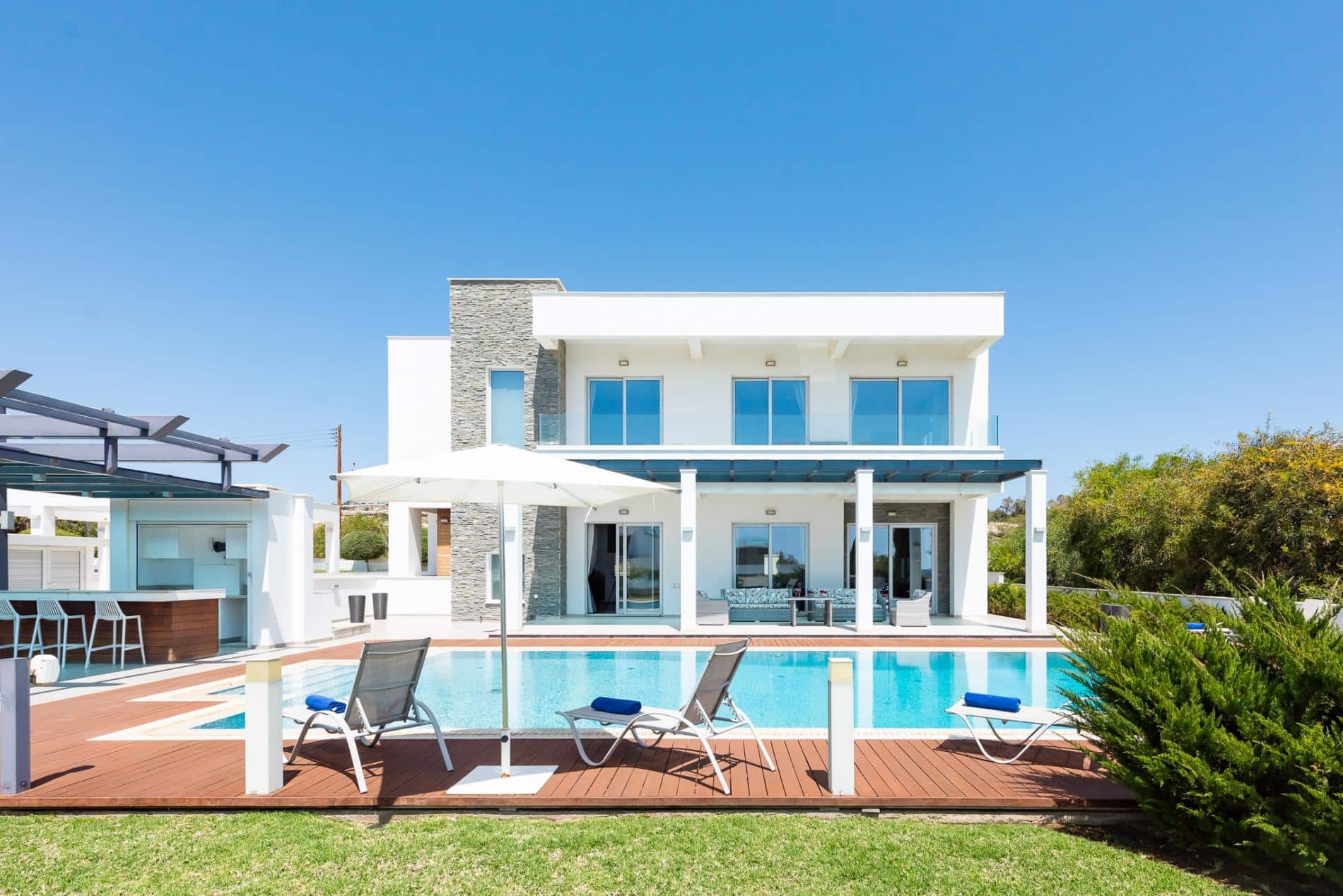 The front of one of Ezoria’a Ayia Napa villas, with garden, wooden pool deck with sunbeds and umbrella around the pool.