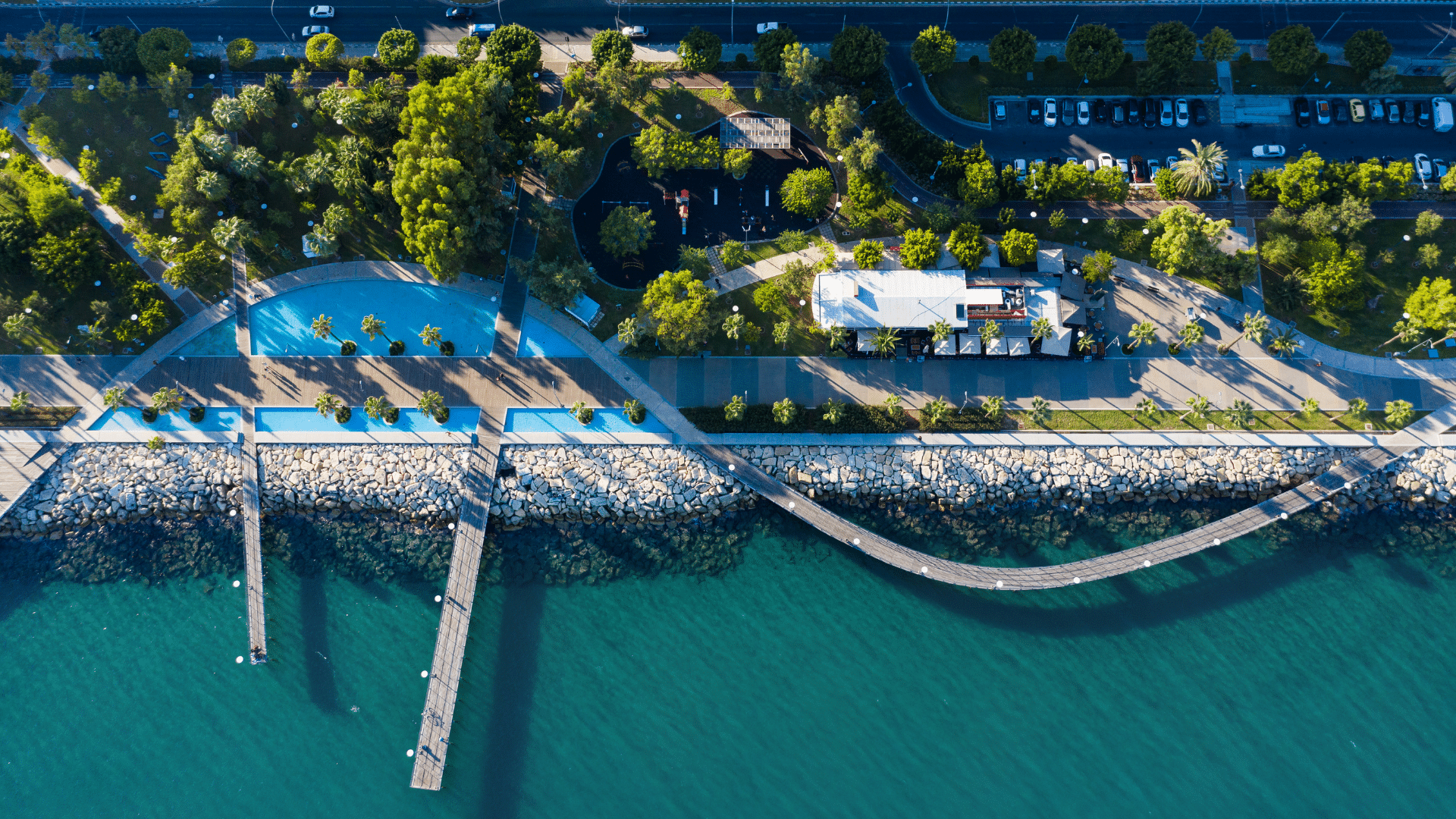 A birds eye view of a walkway, with the sea on one side and a pool and trees on the other.